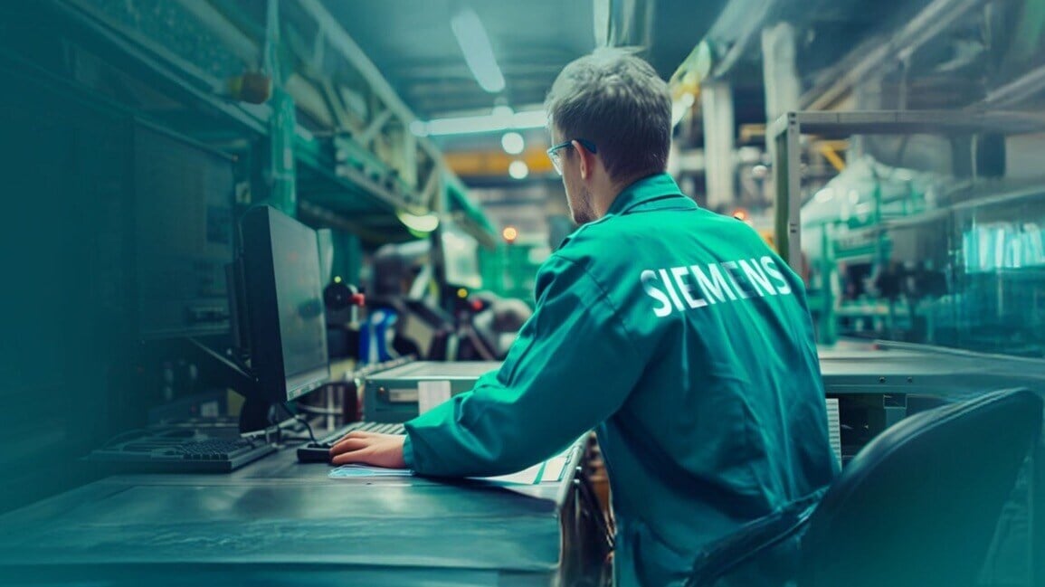A technician in a green Siemens jacket sits in front of a computer in a factory hall with industrial equipment in the background.