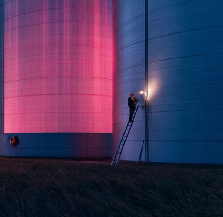 Person on a ladder next to a row of large silos in a field, lit in red and blue at dusk