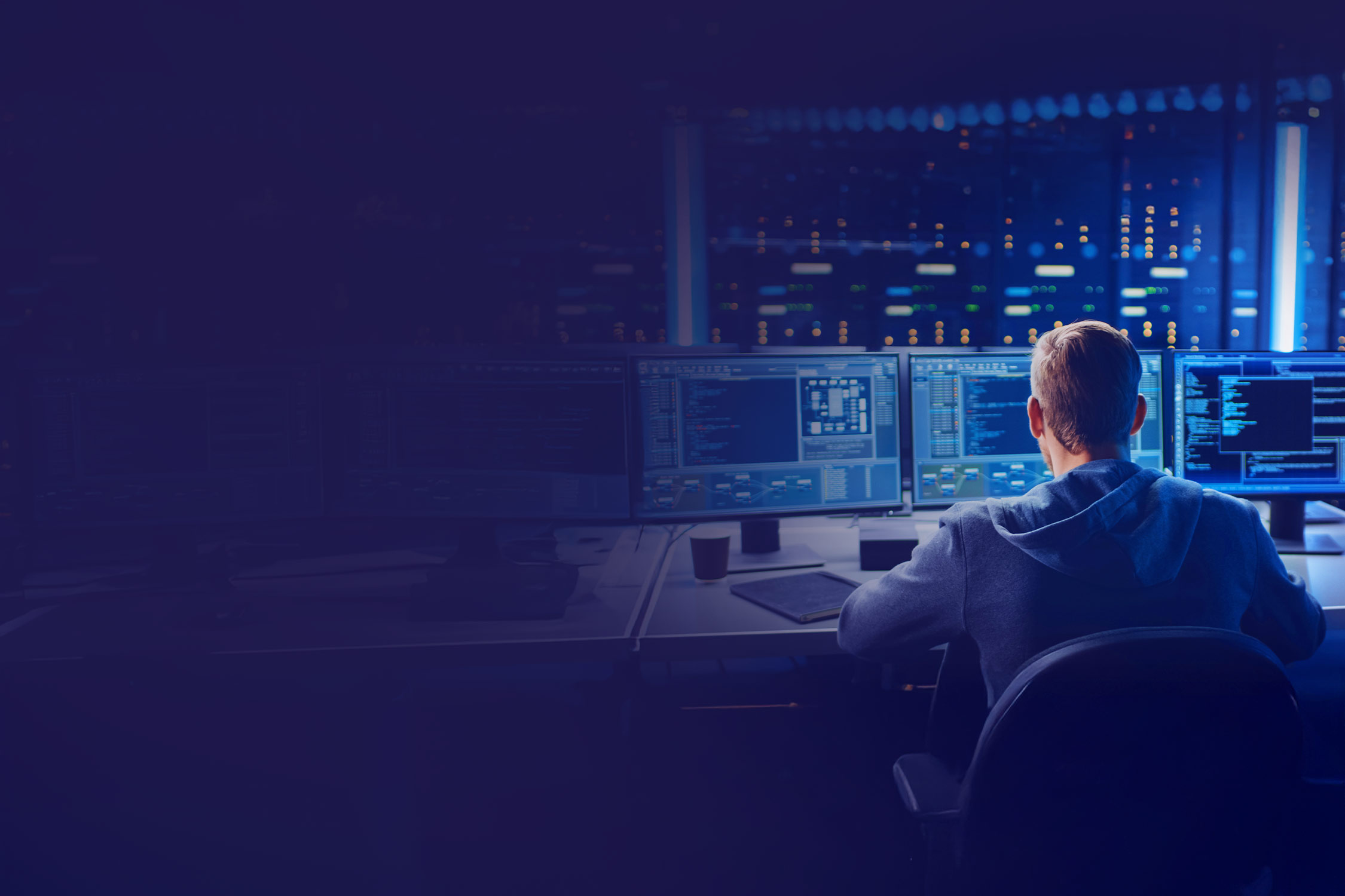 A person works in a dark room at several computer screens showing complex data and programs.