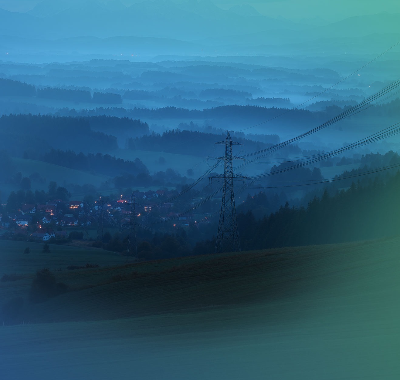 Foggy evening landscape with an illuminated village in the valley, high-voltage pylon and wooded hills in shades of blue and green.