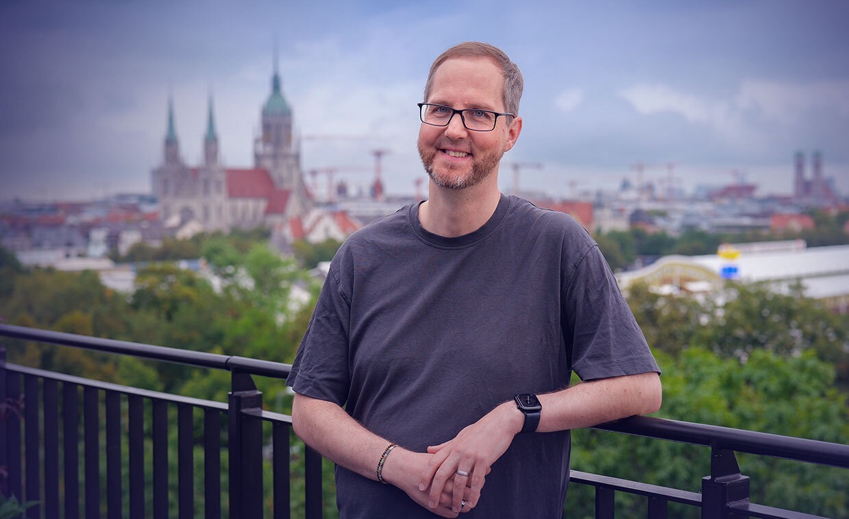 A person wearing a gray T-shirt smiles at the camera. In the background is a panoramic view of Munich.