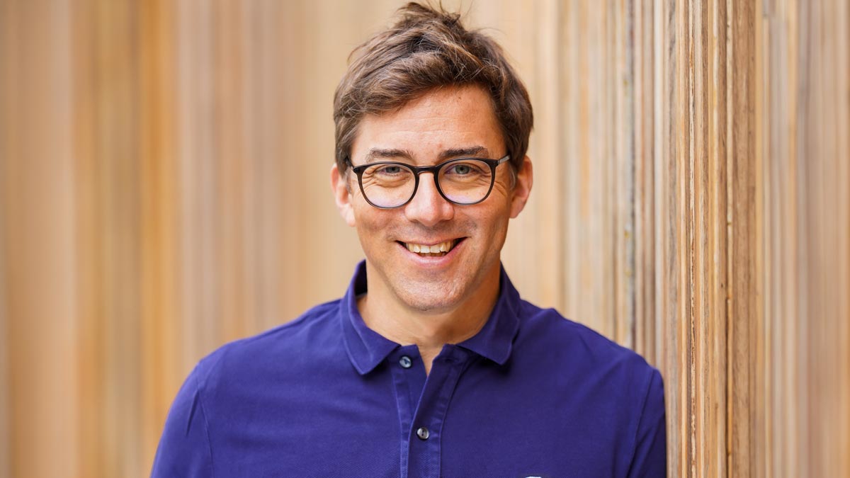 Porträt einer lächelnden Person mit Brille und blauem Poloshirt vor einer hellen Holzwand