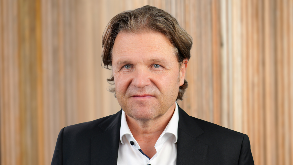 Porträt einer lächelnden Person mit halblangen Haaren und weißem Hemd vor einer hellen Holzwand