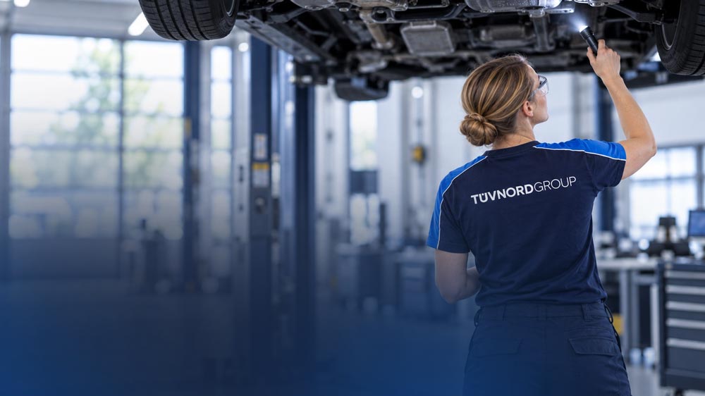 Person in TÜV-Nord-Arbeitskleidung leuchtet mit einer Taschenlampe unter ein aufgebocktes Fahrzeug in einer Werkstatt