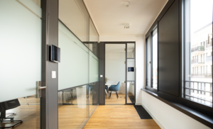 Hallway in the direction of a meeting room. There are glass walls on the left, windows on the right.