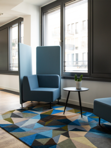 Blauer Sessel mit hohem Sichtschutz, daneben ein kleiner runder Beistelltisch. beides steht auf einem Teppich mit geometrischen Formen, in Herbstfarben gehalten. Im Hintergrund sieht man hohe Fenster. 