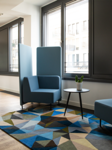 Blue armchair with high privacy screen, next to it a small round side table. both stands on a carpet with geometric shapes, decorated in autumn colors. In the background you can see high windows. 