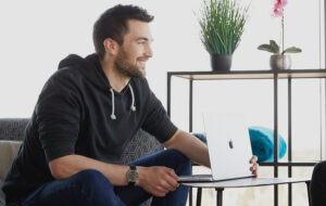 Mitarbeiter im schwarzen Pullover sitzt auf einem Sofa vor einem halb aufgeklappten Macbook