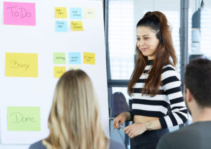 Mitarbeitende vor einem Whiteboard mit bunten Karten darauf, eine Frau steht neben dem Whiteboard, zwei Personen sitzen