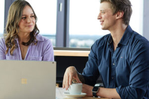 zwei junge Mitarbeitende, eine Frau im lila T-Shirt, ein mann im blauen Shirt, sitzen vor einem Laptop