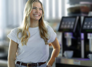 junge Frau, blond, steht vor Kaffeemaschinen