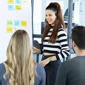 Eine Kollegin erklärt einem Kollegen und einer weiteren Kollegin den Aufbau eines Kanban-Boards, vor dem sie steht. 