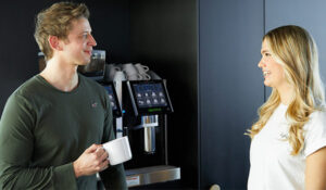Eine Kollegin unterhält sich mit einem Kollegen, der eine weiße Tasse in der Hand hält. Im Hintergrund ist eine Kaffeemaschine zu erkennen.