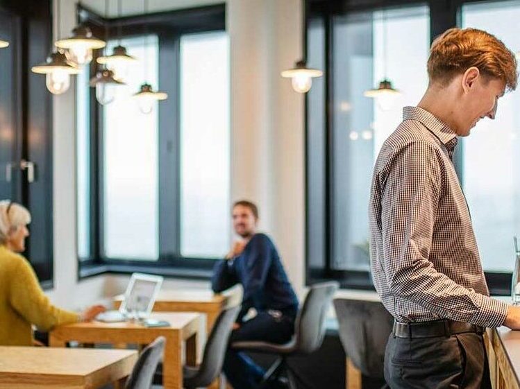 A colleague is standing at a counter, laughing. In the background, a female colleague and a male colleague can be seen, also laughing, in a blur. The scene takes place in our Hamburg office.