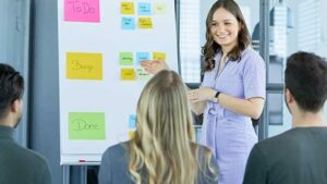 Young woman with brown hair shows two male colleagues and one female colleague a Kanban board that is decorated with colorful pieces of paper. 