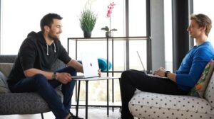 Two young colleagues are sitting across from each other on comfortable armchairs, both with their laptops in front of them.