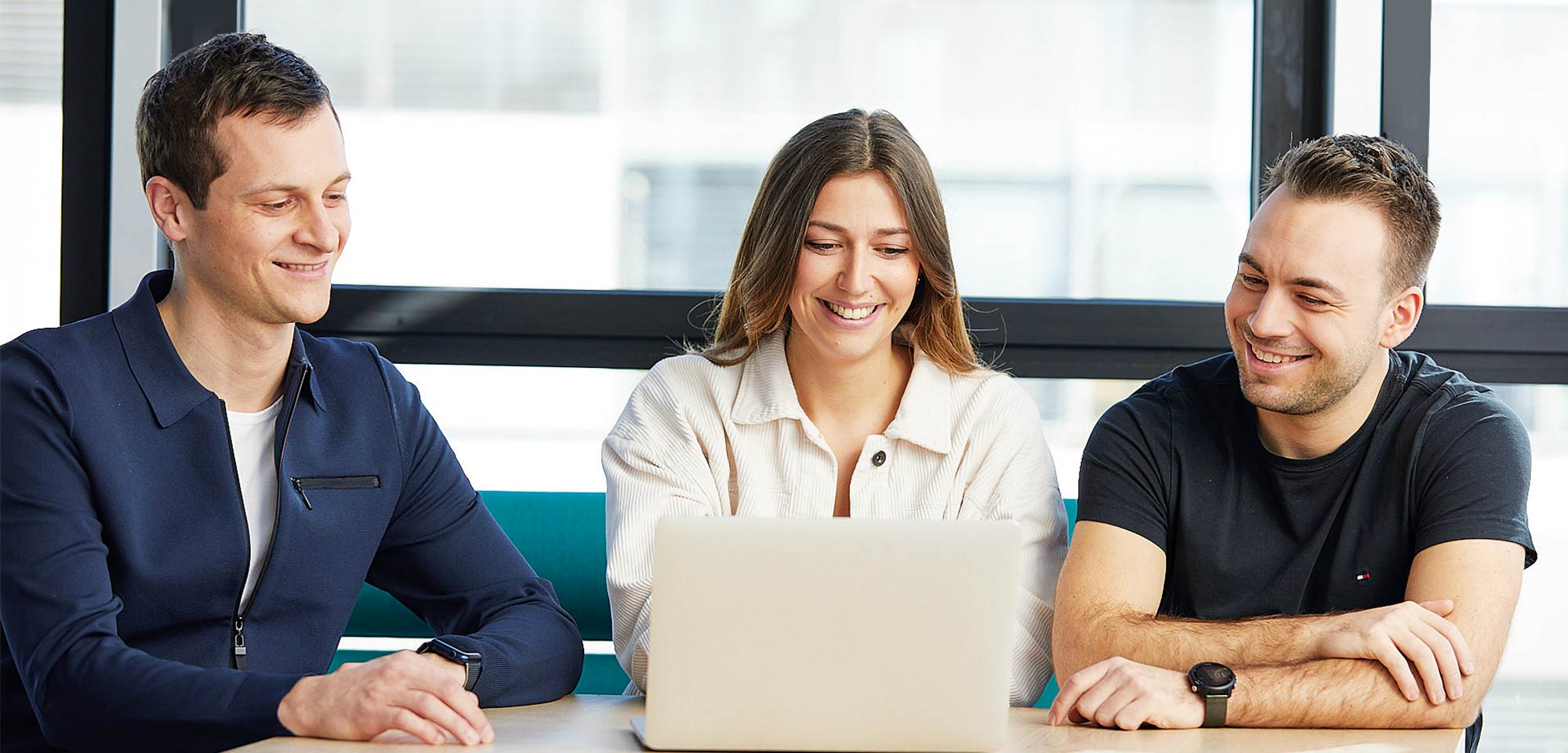 Three colleagues are sitting together at a table, happily looking into a laptop.